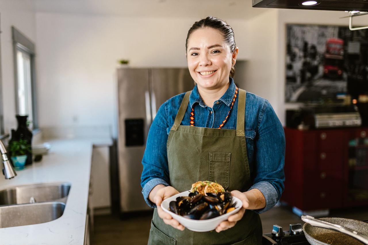 A cheerful woman holding a bowl of cooked mussels in a contemporary kitchen setting.